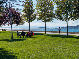 Obraz na płótnie Canvas Istanbul / Turkey - 10.1.2020; New normal: A green park with trees and sea view on a sunny day. A family sits together keeping distance and wearing medical face masks to protect against Covid-19