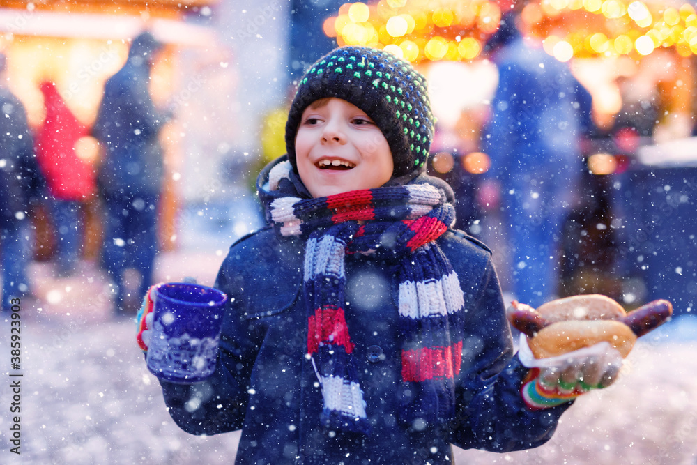 Wall mural little cute kid boy eating german sausage and drinking hot children punch on christmas market. happy