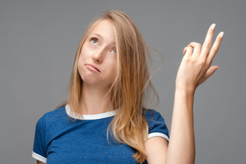 Perplexed blonde girl holding symbolic gun with hand gesture. Studio shot, gray background. Human emotions concept