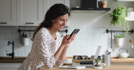 Smiling millennial 35s woman standing in kitchen holding mobile phone looks at screen scroll photos in social media network, spend time on internet websites, choose goods in fashion e-stores at home. - Powered by Adobe