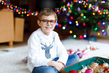 Beautiful kid boy with eye glasses and colorful vintage xmas toys and balls in old suitcase. Child decorating Christmas tree