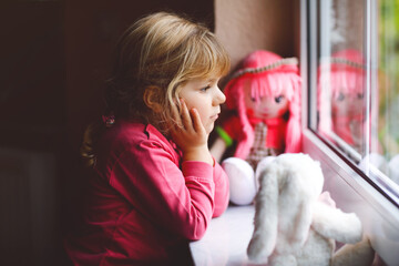 Cute toddler girl sitting by window and looking out on rainy day. Dreaming child with doll and soft...