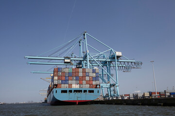 Fototapeta premium Rotterdam, The Netherlands. View of a large containership docked in the container terminal loading and unloading containers on land and river barge moored next to the containership