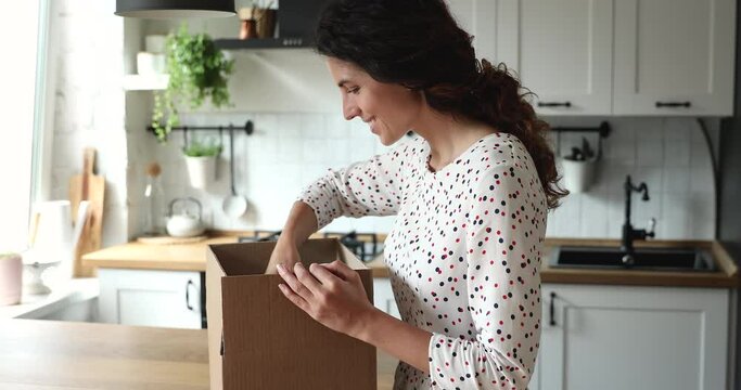 Woman received gift from foreign friend by mail, stands in kitchen opens parcel box looks inside examine carefully ordered high quality goods feels glad. Safe secure express delivery services concept