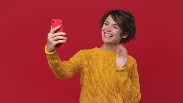 Young fun woman 20s in yellow sweater get video call using mobile cellphone doing selfie videoconference conducting pleasant conversation isolated on red background in studio. People lifestyle concept