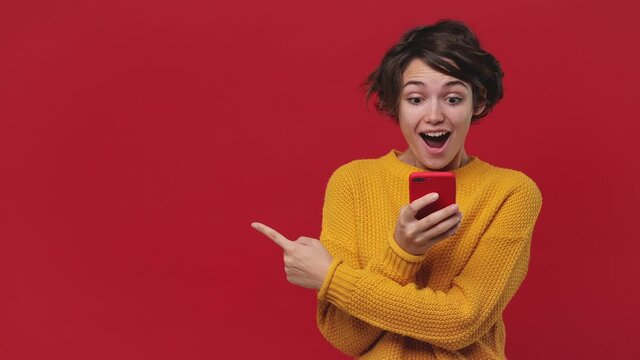 Young fun woman 20s in yellow sweater say wow hold in hand using mobile cell phone pointing fingers aside on workspace mock up copy space isolated on red background in studio. People lifestyle concept