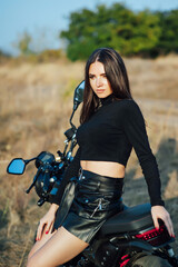 Fototapeta na wymiar Beautiful girl in black clothes sits on motorcycle and poses for camera. Evening sunset