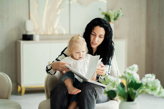 A Brunette Woman Of European Appearance Reads A New Book To Her Little Daughter Baby Kids, An Interesting Fairy Tale With A Happy Ending. Flowers In The Foreground.