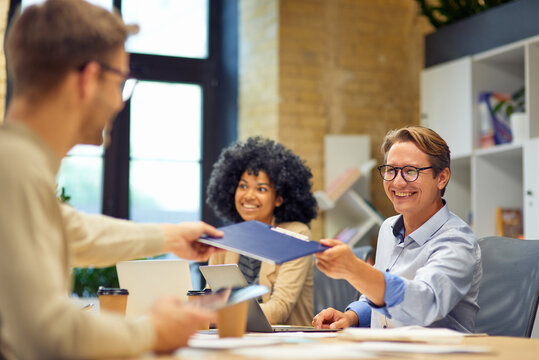 Cheerful man giving a folder with documents to his colleague and smiling while working together in the modern office