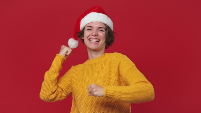 Overjoyed excited shocked fun young woman 20s in yellow cozy sweater Santa Christmas hat doing winner gesture say Yes isolated on red background studi. Happy New Year celebration merry holiday concept