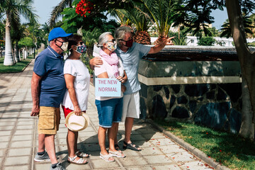 Two couples of mature and senior people wearing surgical mask due to coronavirus Covid-19 outdoor in public park holding message of new normality - concept of active elderly in vacation