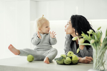 Fototapeta premium girl kids baby blonde and mom brunette, young family playing smiling happiness in the kitchen. holding a large green Apple. modern bright kitchen design. little sweet daughter