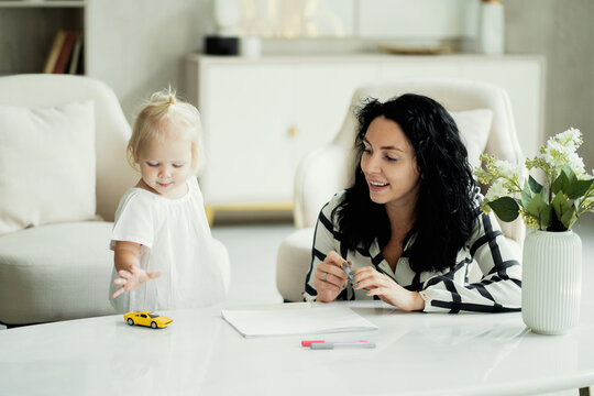 little cute girl blonde European appearance baby and mom brunette kids draws pictures on paper. she was wearing a white shirt and blue jeans. white apartment design, daylight in the window.