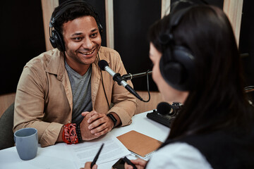 Portait of happy male radio host smiling, talking to female guest while moderating a live show in...