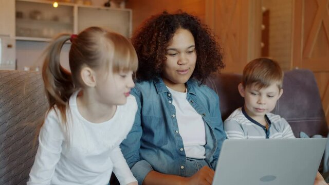 Slow motion of friendly African American nanny talking to children boy and girl and using modern laptop at home. Technology and childcare concept.