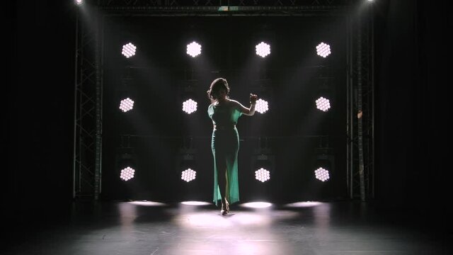 A posh woman in a long sexy dress with a deep slit walks in a dark studio against a background of neon lights. A brunette in high heeled sandals walks straight and looks down. Silhouette. Slow motion.