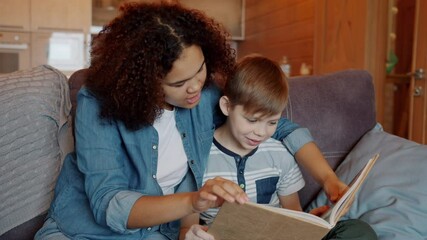 Slow motion of caring Afro-American babysitter reading book to smart kid sitting on sofa at home. Education for preschoolers and babysitting concept.