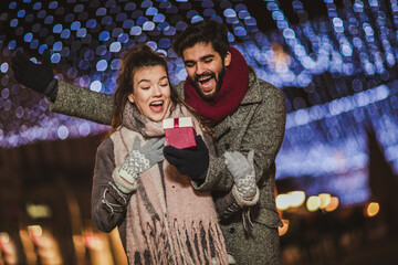 Young couple outdoor with holiday's brights in background. Man presenting gift to woman.