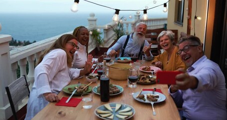 Senior people making video call with smartphone outdoor - Mature friends having fun at dinner night - Slow motion - Powered by Adobe