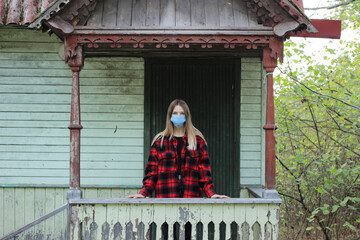 A young blonde woman in a red shirt, self-isolating, stands on the threshold of an old wooden house.
