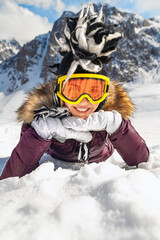 Portrait of playful hipster woman lying on snow at mountain in sunny day with blue sky and big rock in background.