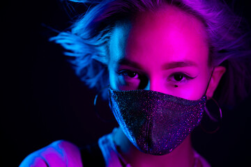 Young beautiful fashionable woman in protective mask with rhinestones dancing in night club. Neon colorful light. Close-up portrait of fashion model.