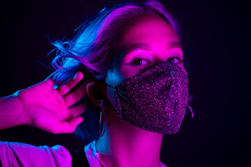 Young beautiful fashionable woman in protective mask with rhinestones dancing in night club. Neon colorful light. Close-up portrait of fashion model.