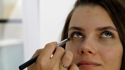 Attractive female face of a brunette well-groomed woman. Makeup artist applies eye shadows to the eyelid of the model eyes. Portrait of a beautiful young woman close up. Professional stylish makeup. 