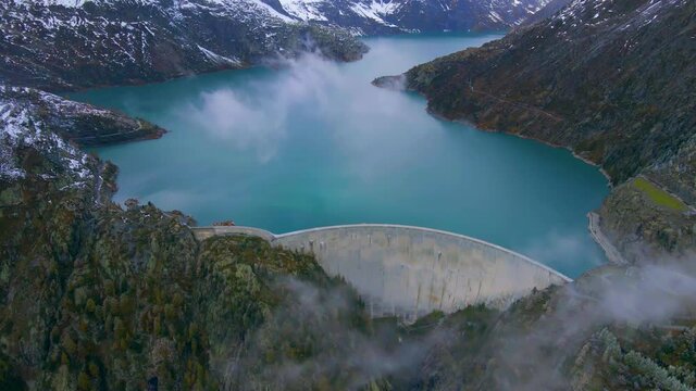 Hydropower generated from arch dam and reservoir lake in snow covered Swiss Alps mountains to produce renewable energy, sustainable hydroelectricity, aerial drone footage 4K video, autumn weather