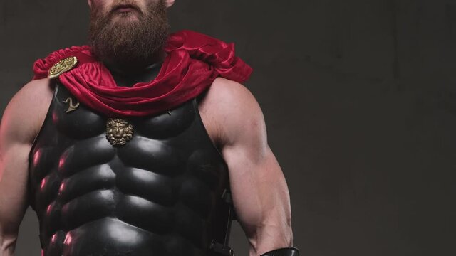 Warlike and bearded rome warrior with black armour and red cape posing while camera rotates and showing his muscular build biceps and abs in dark background in studio.