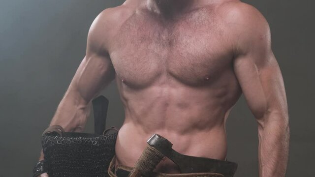 Camera shoots moving up from below showing muscular and naked scandinavian warrior he holds his helmet with hatchet on his belt then viking wears his helmet on head and takes an axe from belt and