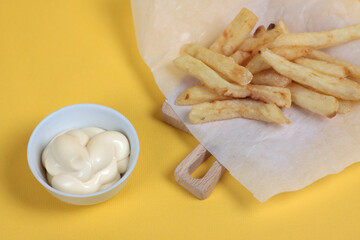 Fototapeta na wymiar a pile of french fries on parchment paper and a bowl of white sauce on cutting board on yellow background flat lay
