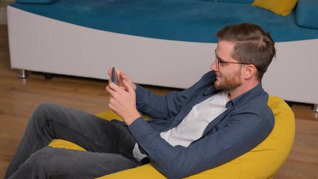 Young Bearded Man Sits On Bean Bag And Playing A Game Using A Phone, At Home. Having Fun.