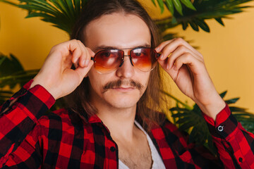 An attractive man with long hair and a mustache in a red plaid 80s disco shirt sits on a chair...
