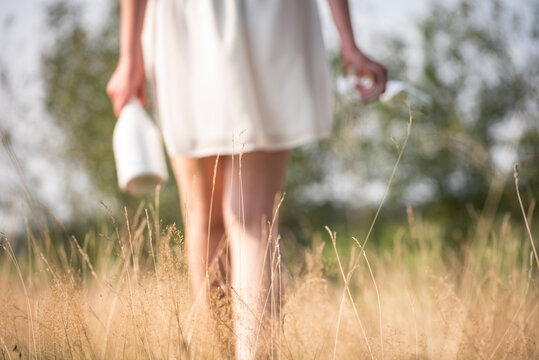 Blurred picture of a young woman with a white dress going through high grass with a bottle of sparkling wine and two glasses in her hand