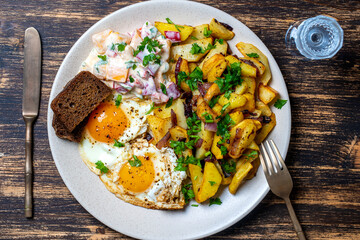 Ukrainian food, fried potatoes with onions, eggs, vegetable salad, black bread and glass of vodka on wooden background