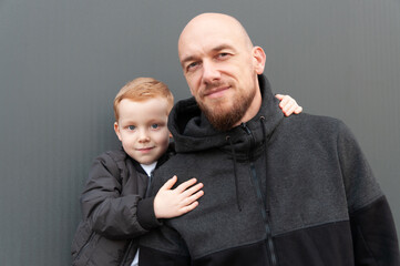 Little red-haired son hugs dad on a gray background, dressed in black and gray clothes