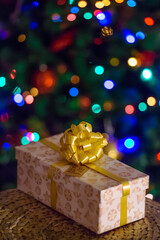 Beautiful Christmas gift with a gold bow under the tree against a background of multi-colored bokeh lights blurry out of focus. Postcard, wallpaper, vertical