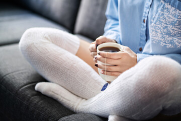 Long cozy warm socks and a hot cup of tea in winter. Woman enjoying relaxed lifestyle on home couch. Sick person with flu or fever sitting on sofa during cold season. Comfortable peaceful weekend.