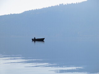fisherman in boat silhouette on calm lake with mountain reflecting on water
