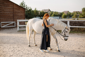 A beautiful red-haired girl in a dress walks with a horse at the horse ranch. Portrait of a girl...