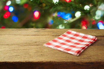 Obraz premium Christmas holiday background with empty wooden table with red checked tablecloth over Christmas tree bokeh. Product montage display or mock up design