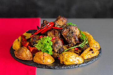 Baked meat with potatoes with skin on a round black plate in the original setting. Grilled steaks, baked potatoes and зелень. 