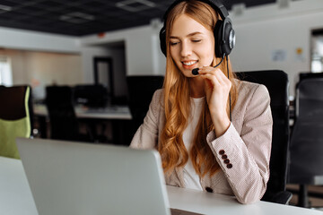Smiling young female operator in headset works while sitting at the table, uses laptop, working in...