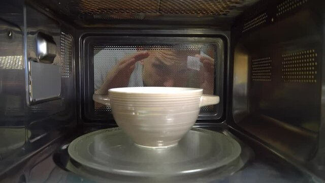 A ceramic bowl with some food is being heated in the microwave, and a man's face looks nervously and discontentedly into the microwave through the glass