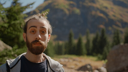 Hipster man with long beard with backpack behind him is looking towards the camera with a confused facial expression and thinks, lifestyle and nature concept