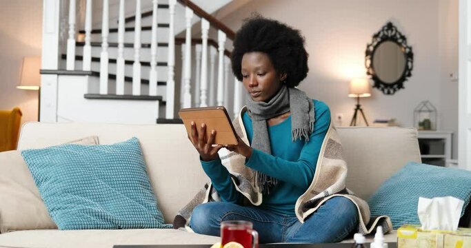 Sick beautiful woman in plaid sitting on sofa and typing on tablet in room. Unwell African American young female tapping and scrolling on device indoors at home. Virus infection concept