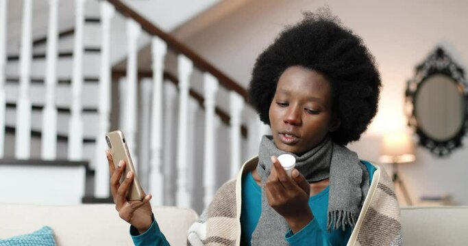 Close up portrait of sick woman in scarf sitting in room and videochatting on cellphone. Unwell African American young female talking on video on smartphone at home and showing pills. Disease concept