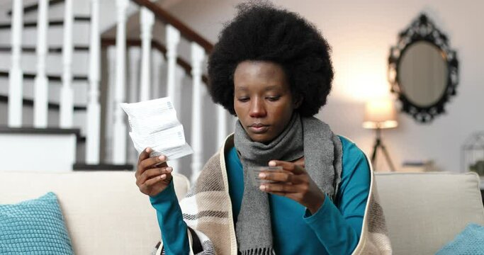 Close up portrait of African American female sitting at home on sofa and reading prospectus for medicine. Sick young woman holding pills in hand in room. Pandemic virus. Disease treatment concept