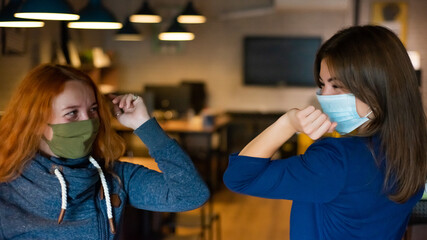 Two caucasian women wearing face masks greet bumping elbows in doors